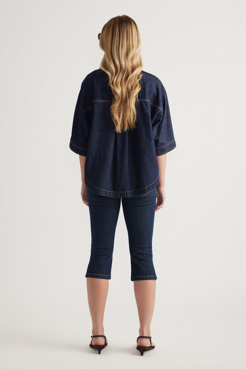 Back view of a woman wearing a relaxed dark denim shirt with cuffed sleeves and cropped jeans
