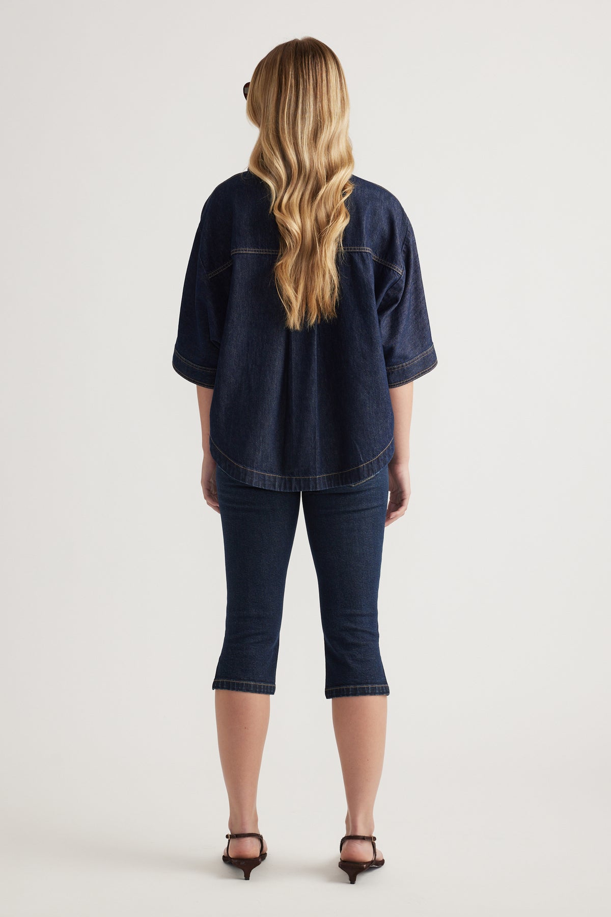 Back view of a woman wearing a relaxed dark denim shirt with cuffed sleeves and cropped jeans