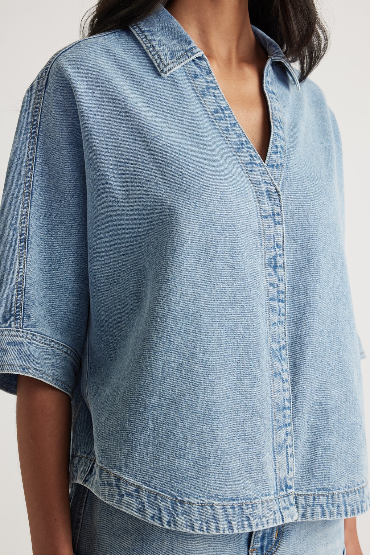 Woman wearing a light wash denim shirt with a collared V neckline, cuffed sleeves and scooped hem