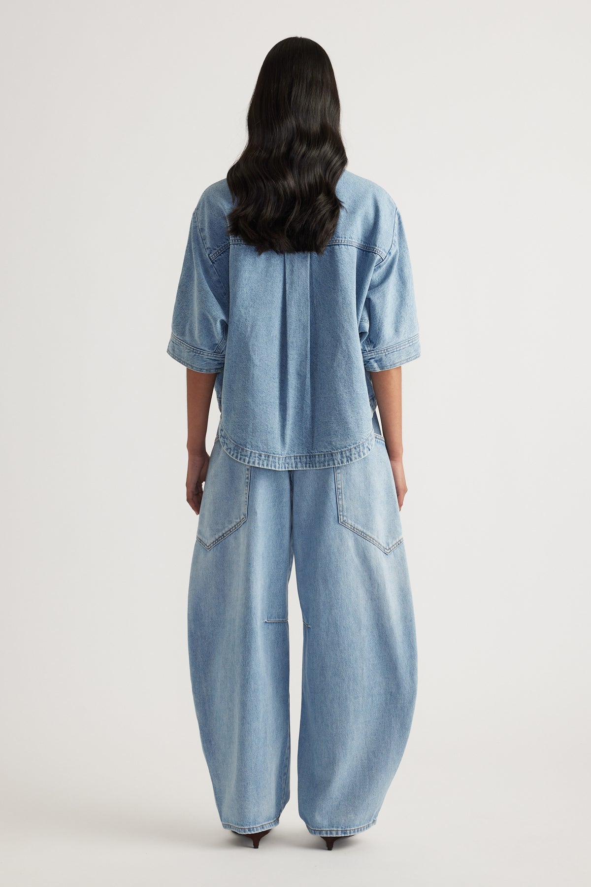 Back view of a woman wearing a light wash denim shirt with cuffed sleeves and matching wide leg jeans
