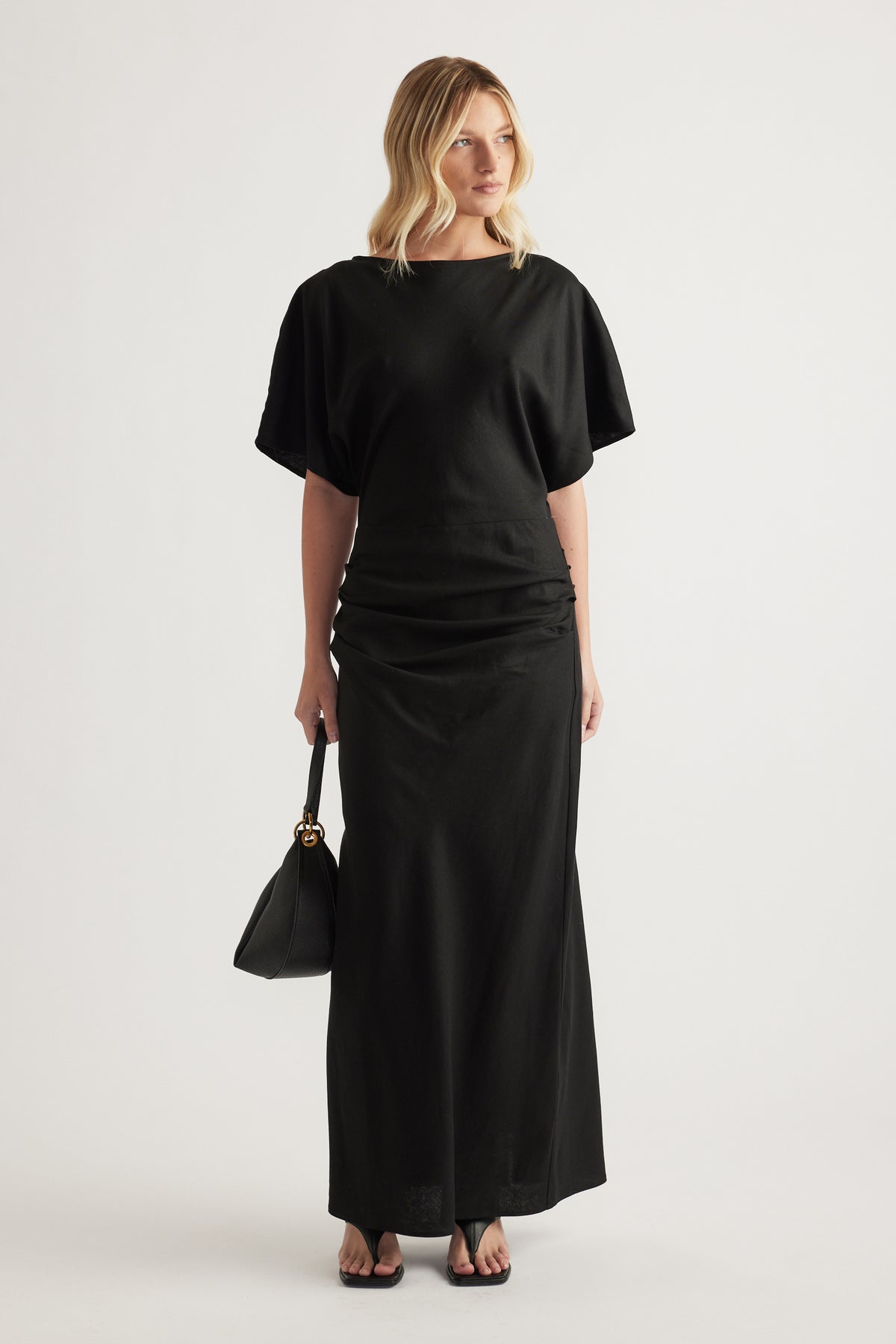 Woman wearing a black boat neck short sleeve ruched maxi dress with a black handbag and sandals