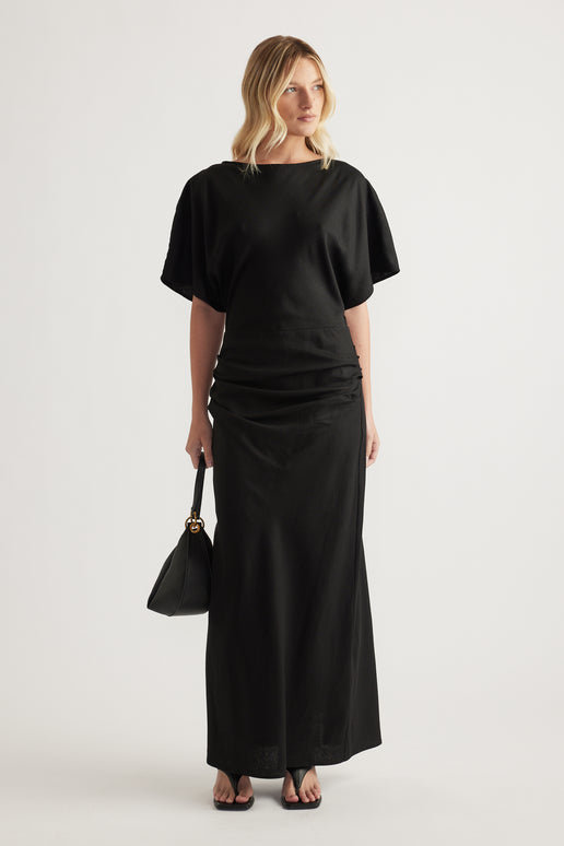Woman wearing a black boat neck short sleeve ruched maxi dress with a black handbag and sandals