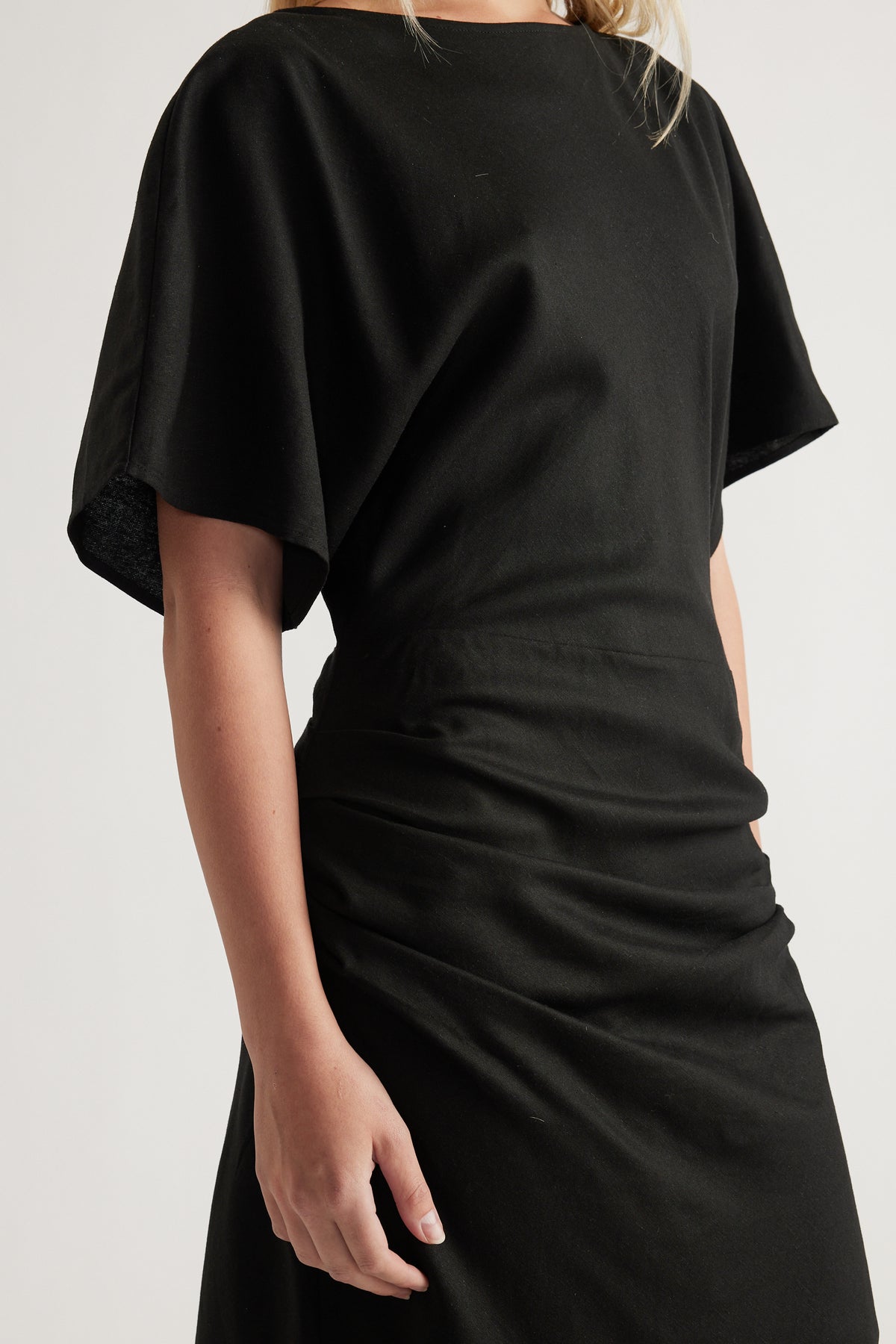 Woman wearing a black boat neck midi dress with short sleeves and ruched detailing at the waist and skirt