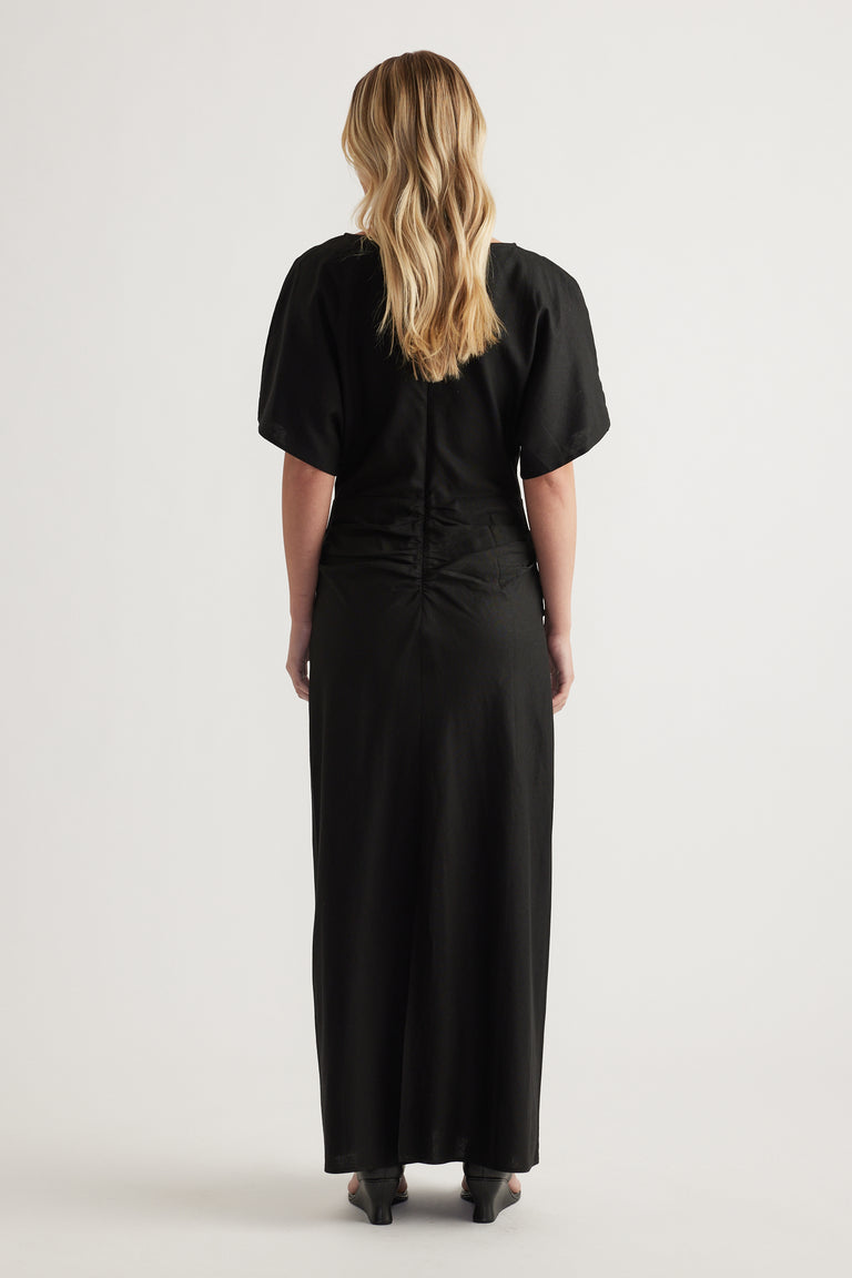 Woman viewed from behind wearing a black short sleeve maxi dress with ruched back detail and a flowing hem