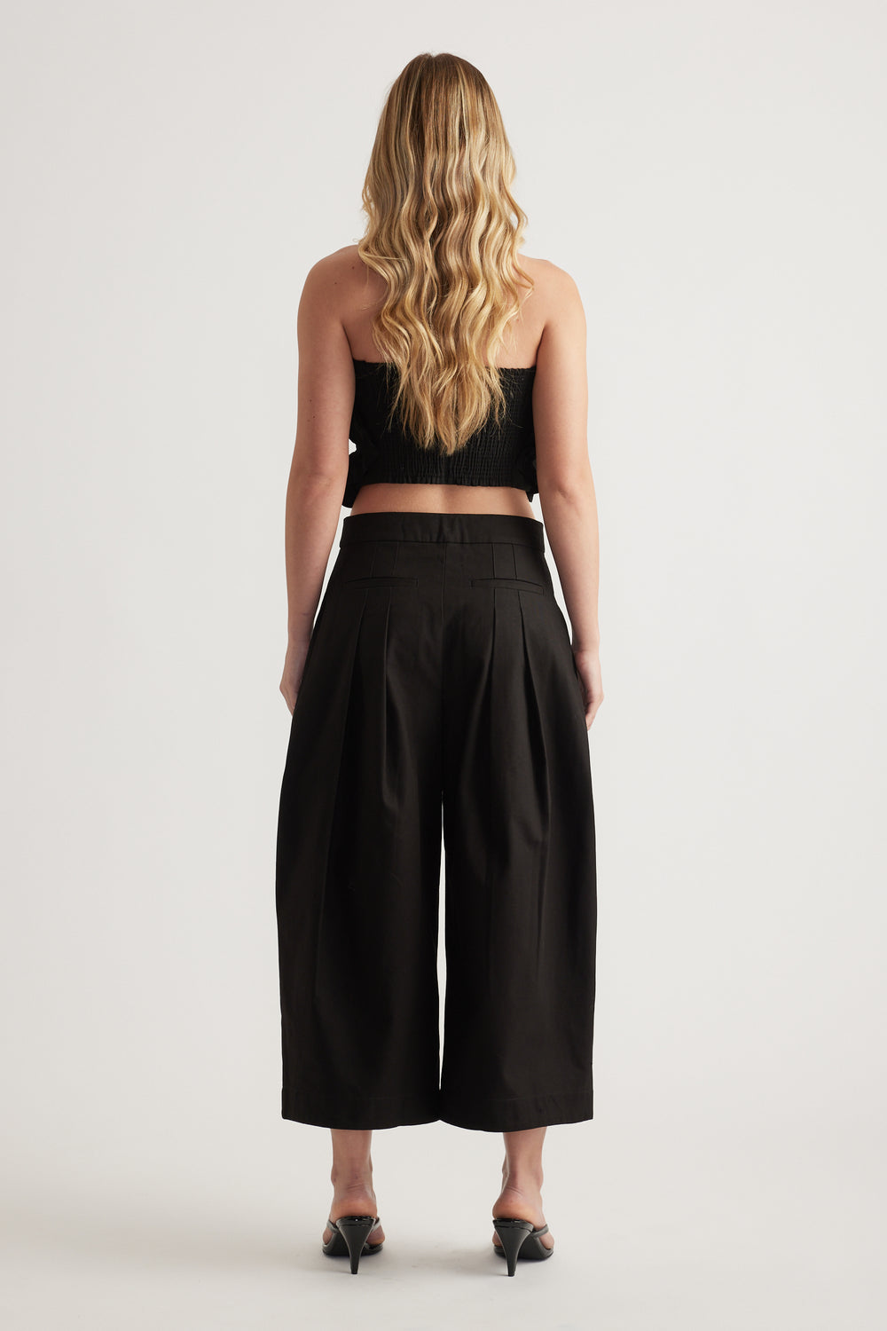 Back view of a woman wearing black pleated cropped wide leg pants with a black top and heels