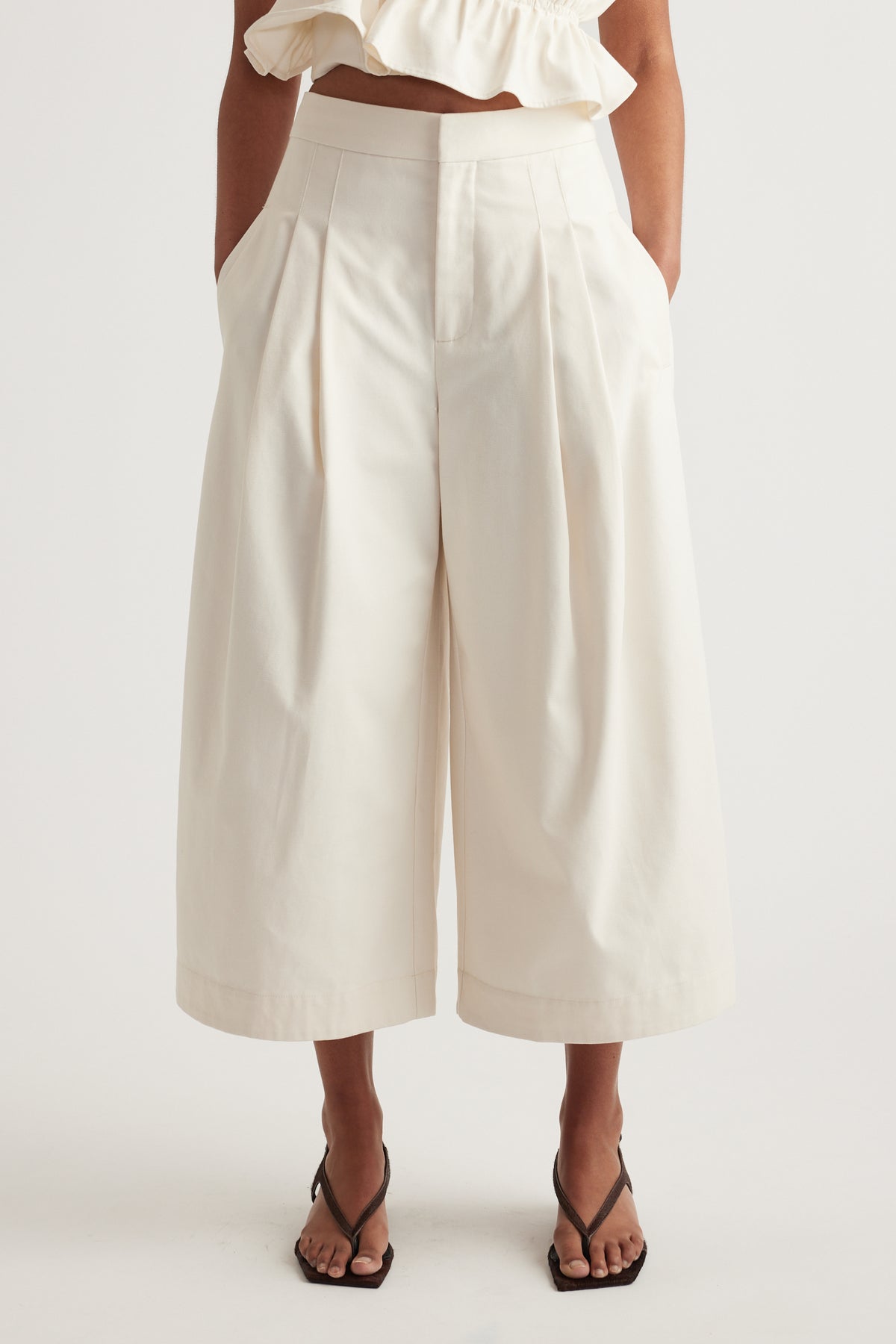 Woman wearing white high rise pleated cropped wide leg pants with a ruffle top and black strappy sandals