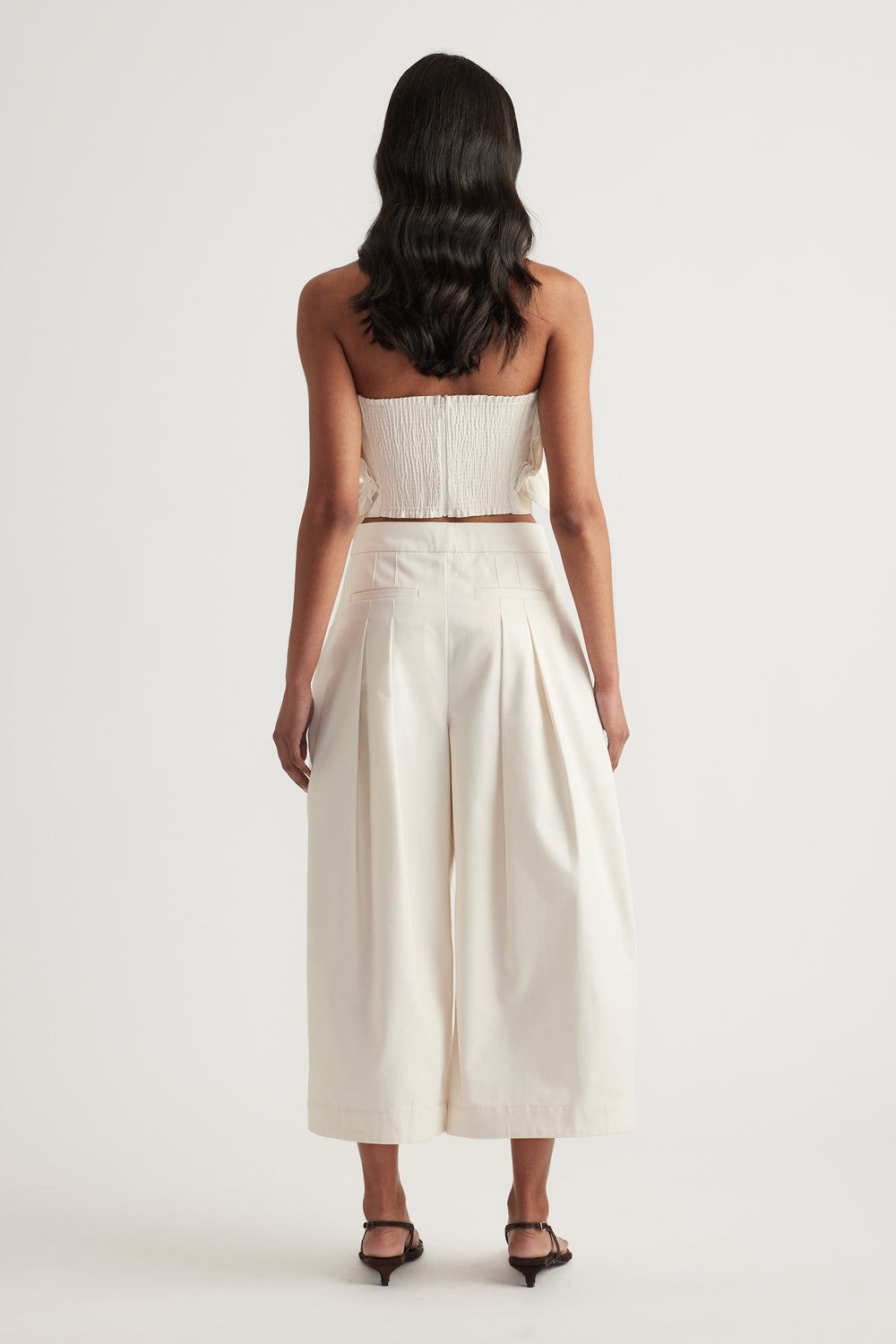 Back view of a woman wearing a cream strapless top with pleated wide leg pants and black heels