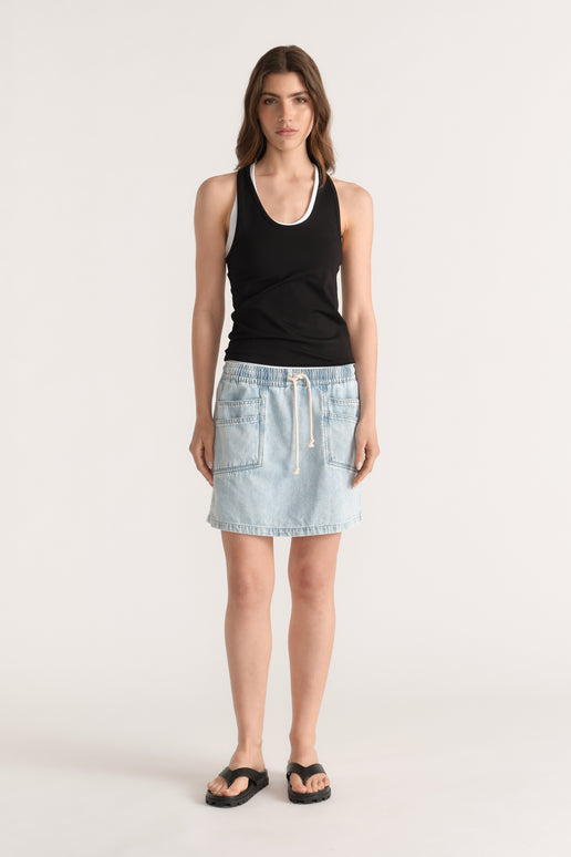 Woman wearing a light wash denim mini skirt with an elastic drawstring waist, black tank top and flat sandals