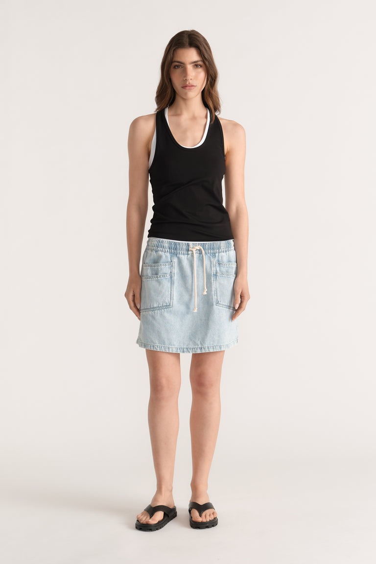 Woman wearing a light wash denim mini skirt with an elastic drawstring waist, black tank top and flat sandals