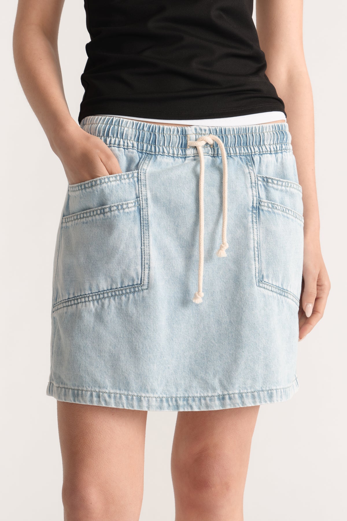 Model wearing a light wash denim mini skirt with an elasticated waistband, tie closure and patch pockets