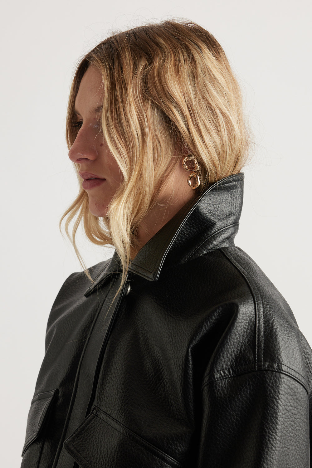 Woman with wavy blonde hair wearing textured shiny gold open drop earrings and a black leather jacket with collar upturned