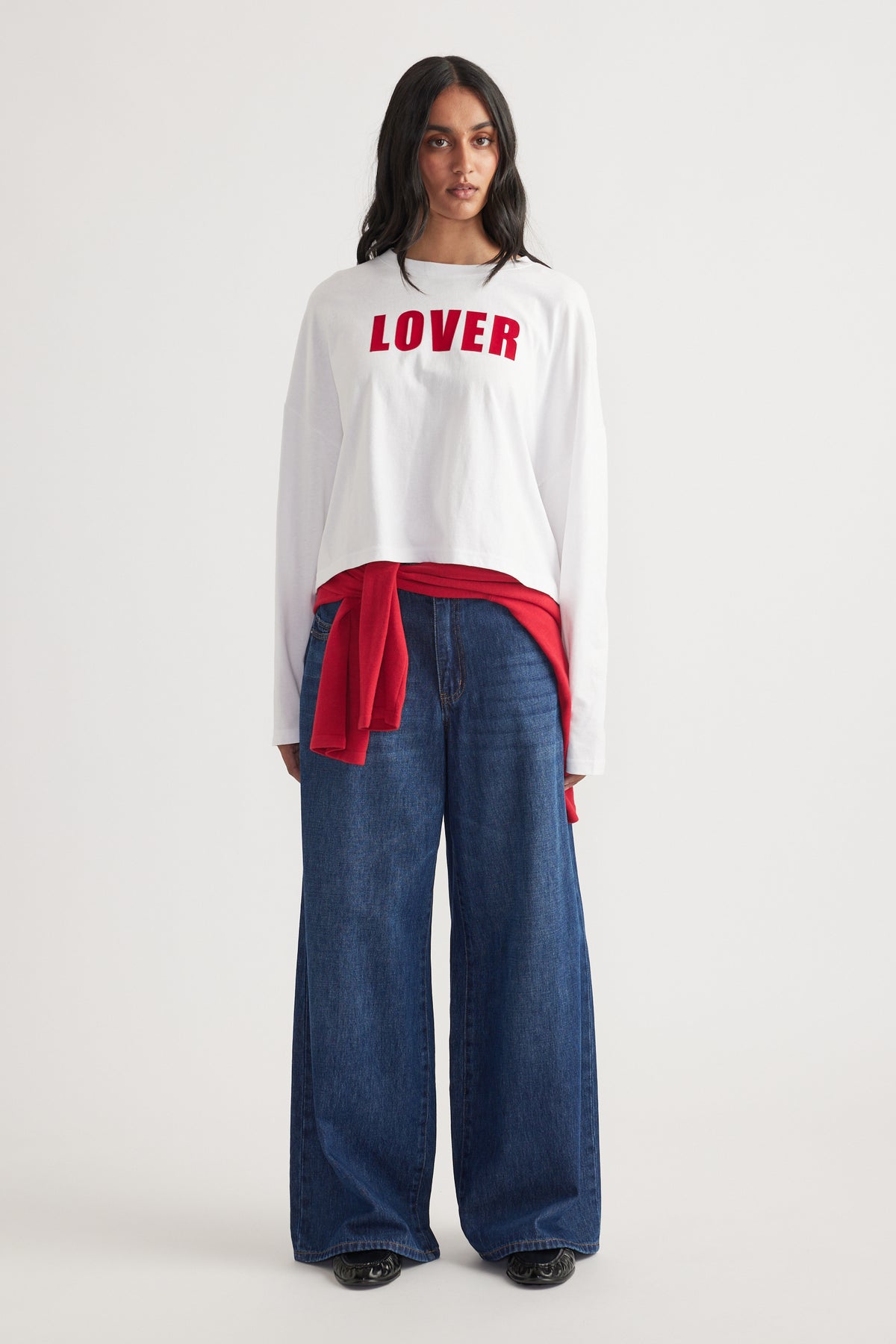 Woman wearing a white long sleeve top with red text, paired with wide-leg dark blue jeans and a red sweater tied at waist