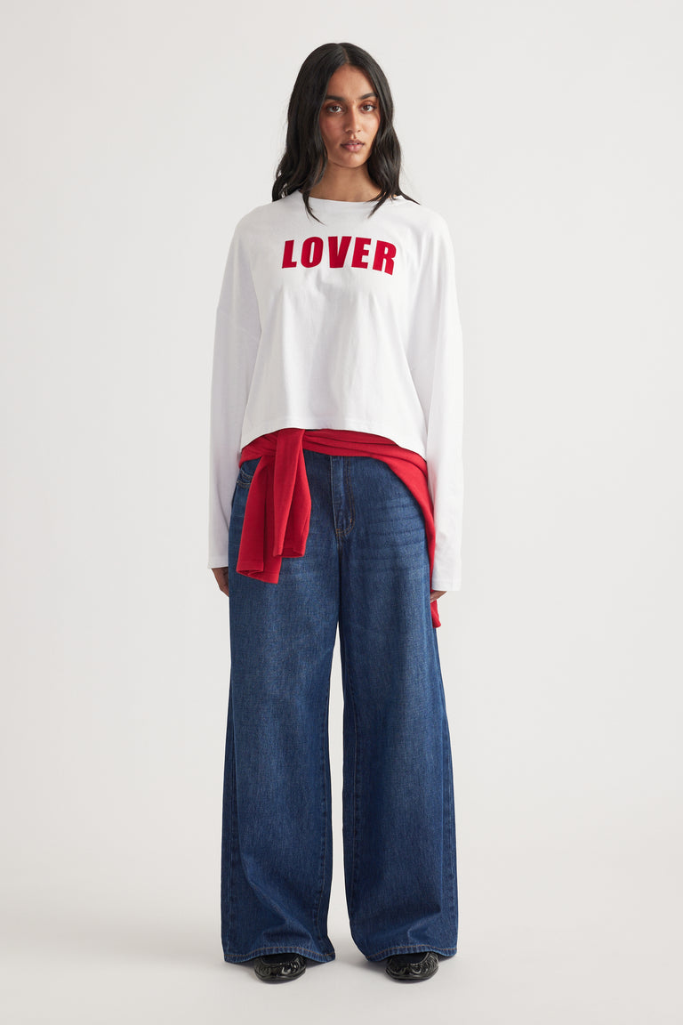 Woman wearing a white long sleeve top with red text, paired with wide-leg dark blue jeans and a red sweater tied at waist