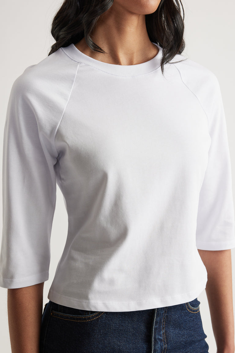 Woman wearing a fitted white crew neck tee with three-quarter raglan sleeves and seam detailing, paired with dark jeans