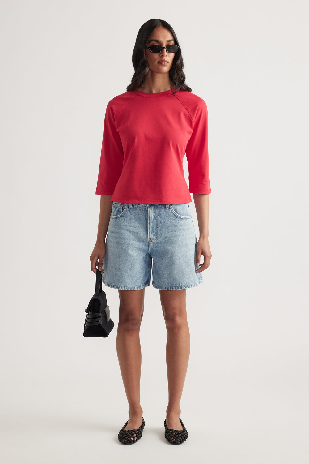 Woman wearing a red fitted three-quarter sleeve tee with denim shorts and sunglasses