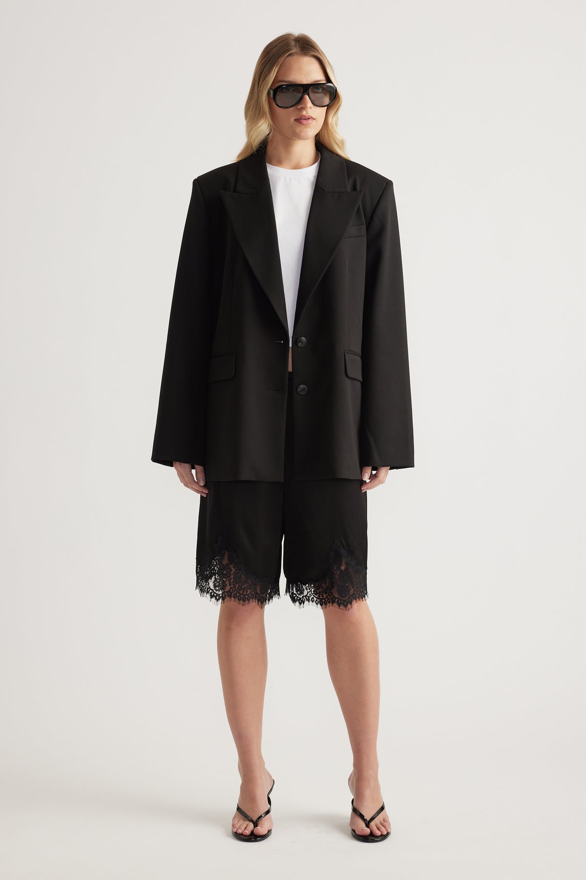 Woman wearing a black blazer, white tee and black lace trim Bermuda shorts with strappy sandals