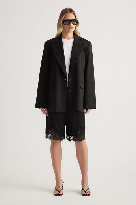 Woman wearing a black blazer, white tee and black lace trim Bermuda shorts with strappy sandals