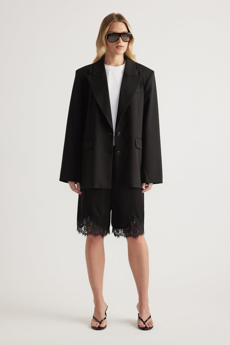 Woman wearing a black blazer, white tee and black lace trim Bermuda shorts with strappy sandals