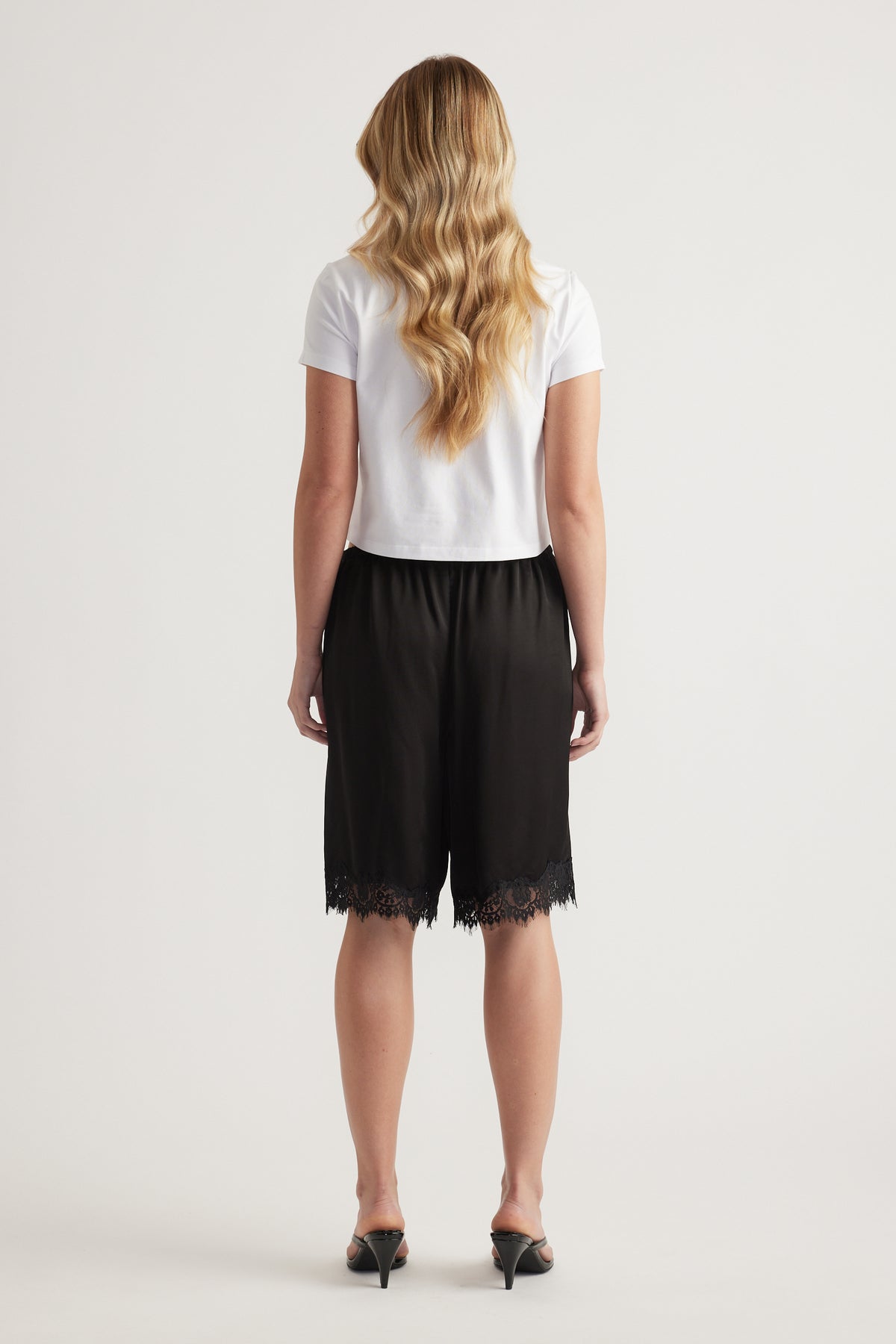 Back view of a woman in a white tee and black satin Bermuda shorts with lace trim and heels