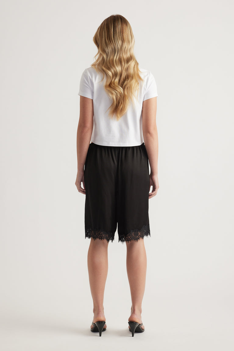 Back view of a woman in a white tee and black satin Bermuda shorts with lace trim and heels