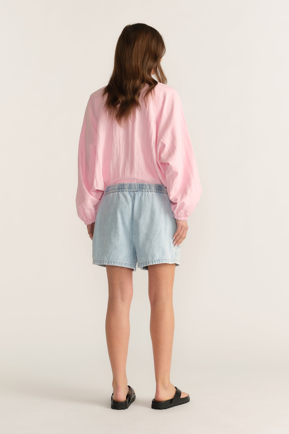 Back view of a woman wearing light wash denim shorts with a pink long sleeve top and black sandals
