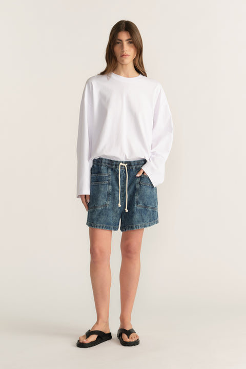 Woman wearing mid rise denim shorts with an elastic drawstring waist, a white long sleeve top and black sandals