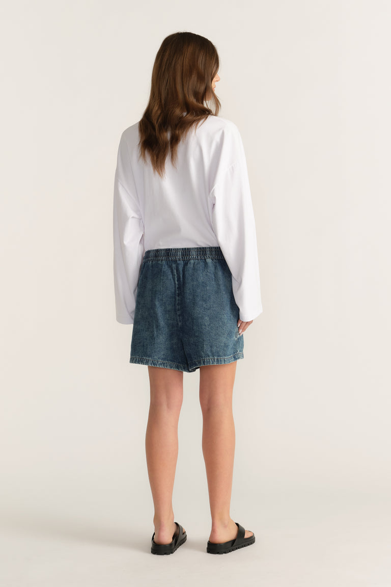 Back view of woman wearing mid-rise denim shorts with a white long-sleeve shirt and black sandals