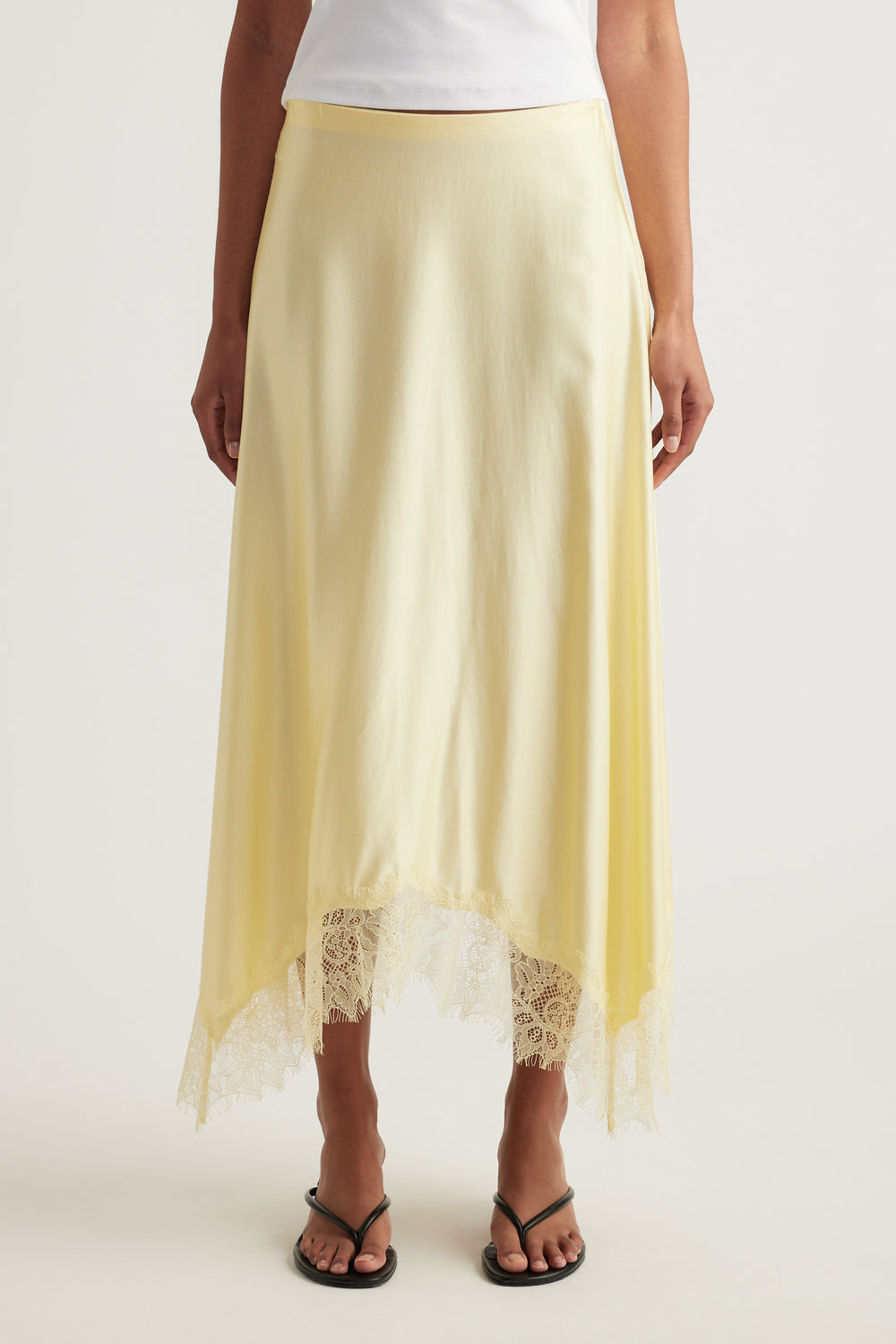 Woman wearing a yellow satin midi skirt with lace trim and handkerchief hem, styled with a white top and sandals