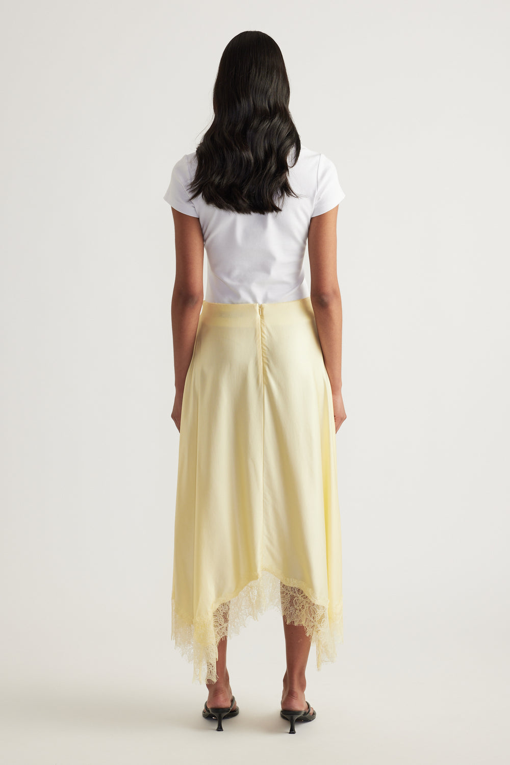 Back view of a woman wearing a pale yellow satin midi skirt with a lace trimmed handkerchief hem and white tee