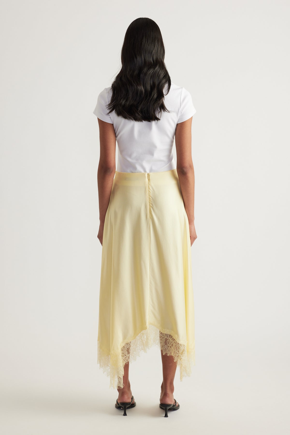 Back view of a woman wearing a pale yellow satin midi skirt with a lace trimmed handkerchief hem and white tee