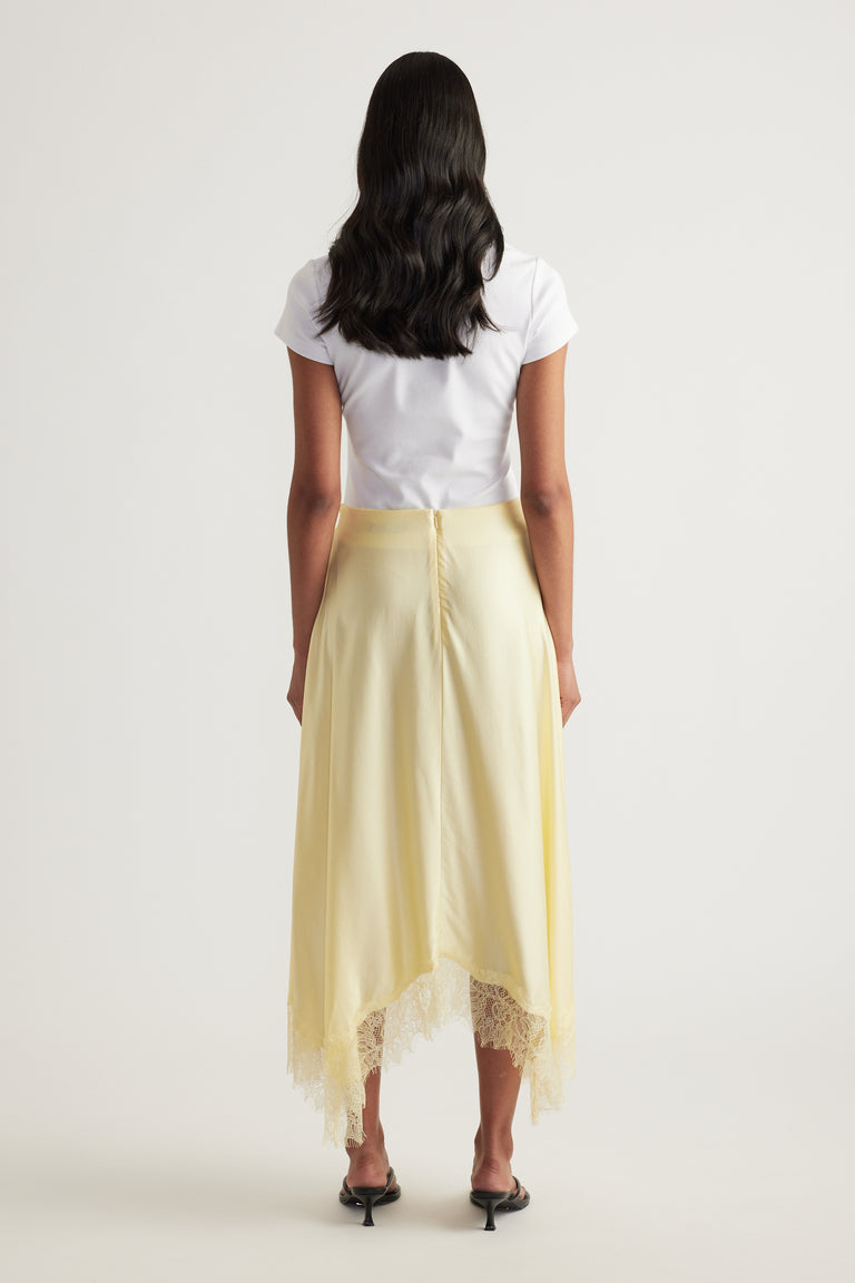 Back view of a woman wearing a pale yellow satin midi skirt with a lace trimmed handkerchief hem and white tee