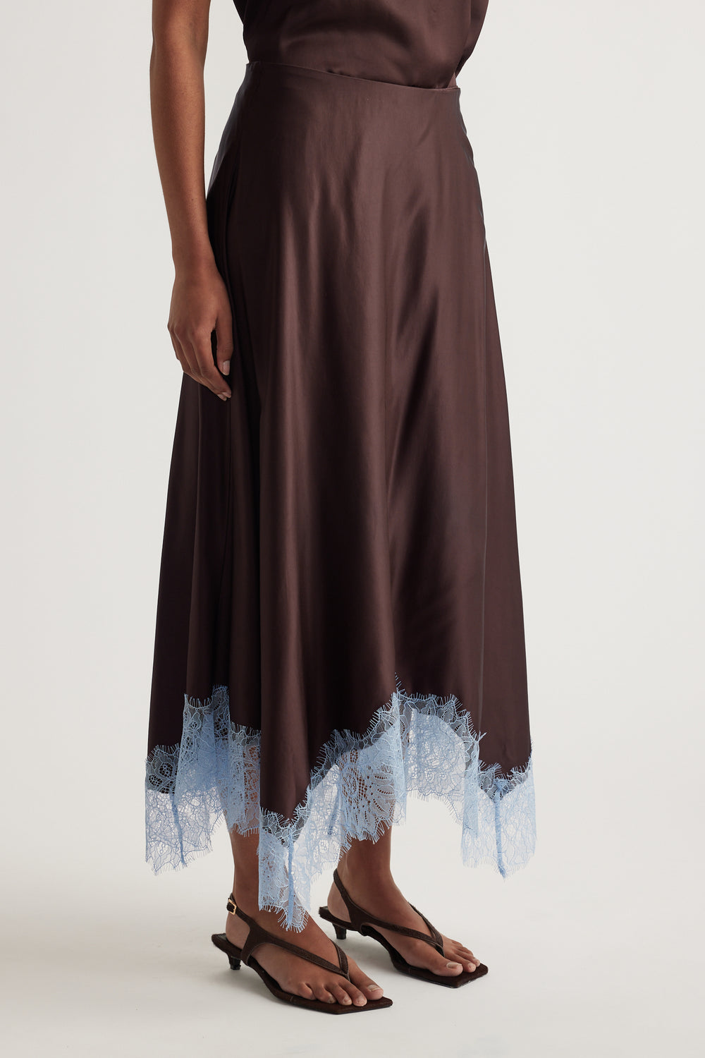 Woman wearing a chocolate brown satin midi skirt with blue lace trim and black strappy heels
