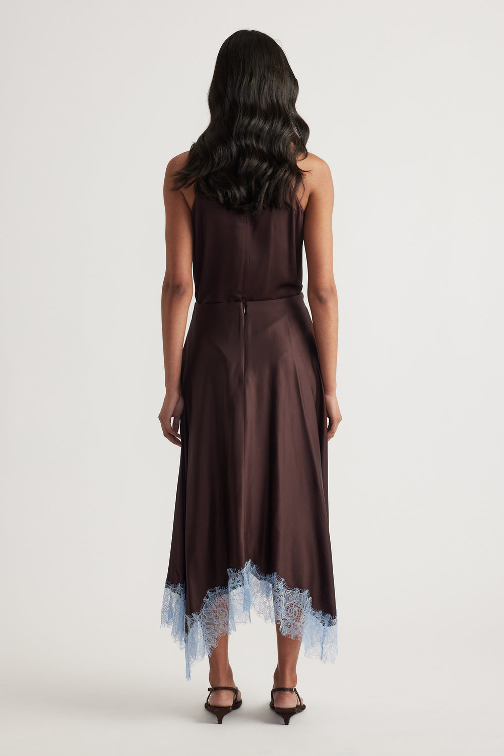 Back view of a woman wearing a chocolate satin midi skirt with blue lace trim and black heels