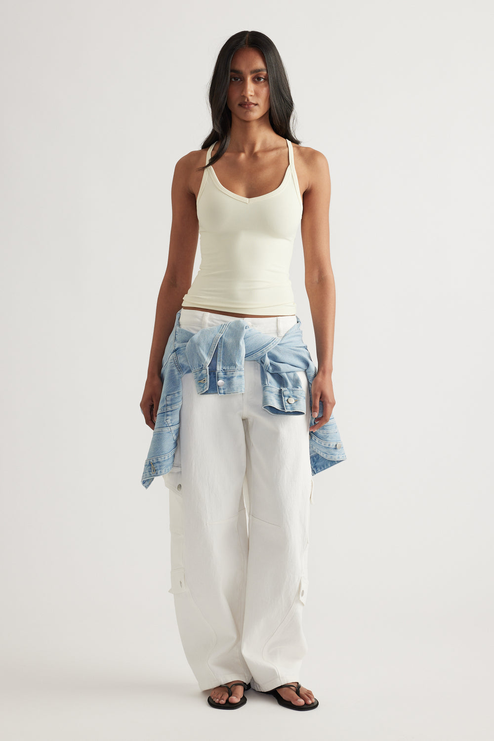 Model wearing a fitted white tank top with white wide leg pants and a denim jacket tied around her waist