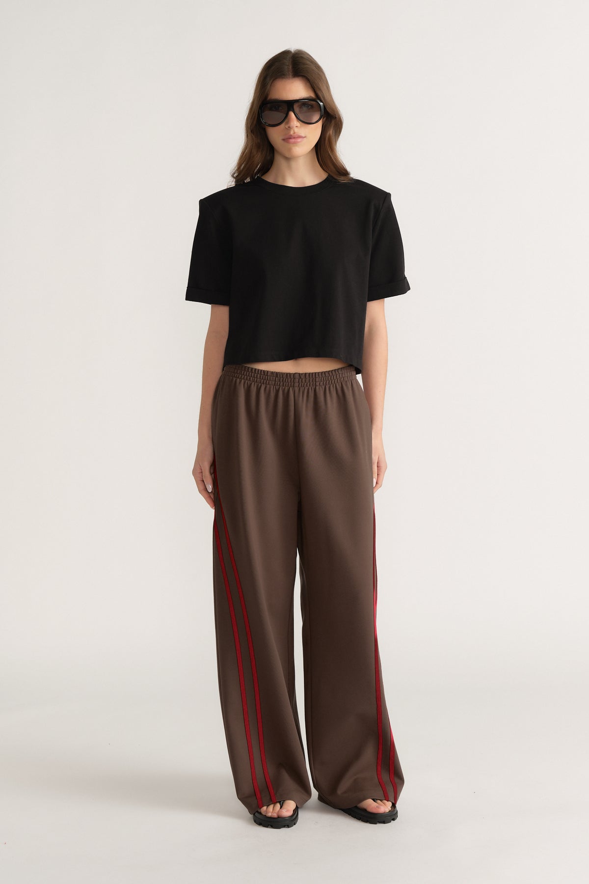 Model wearing a black cropped shoulder pad tee with brown wide leg pants featuring red side stripes and sunglasses