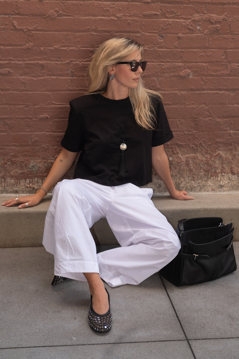 Woman wearing a black padded shoulder tee with white wide leg pants sunglasses and a black handbag
