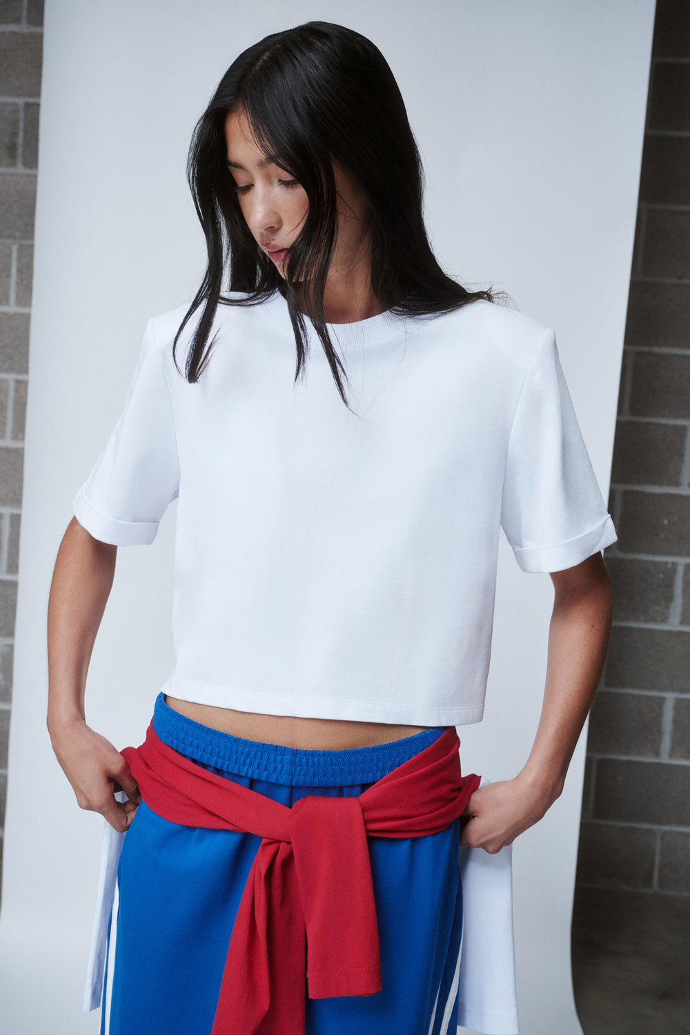 Woman wearing a white shoulder pad tee with blue pants and a red sweater tied at the waist