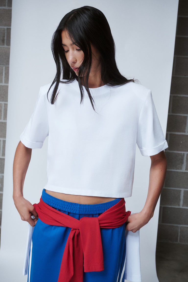 Woman wearing a white shoulder pad tee with blue pants and a red sweater tied at the waist