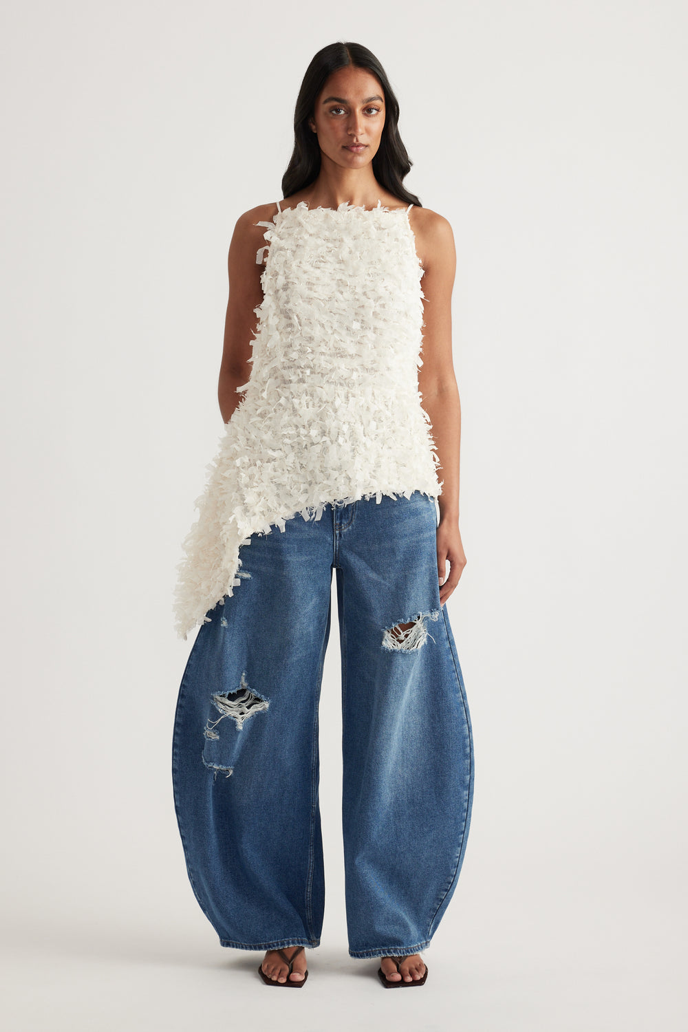 Woman wearing a white textured asymmetrical top with thin straps and wide leg distressed denim jeans
