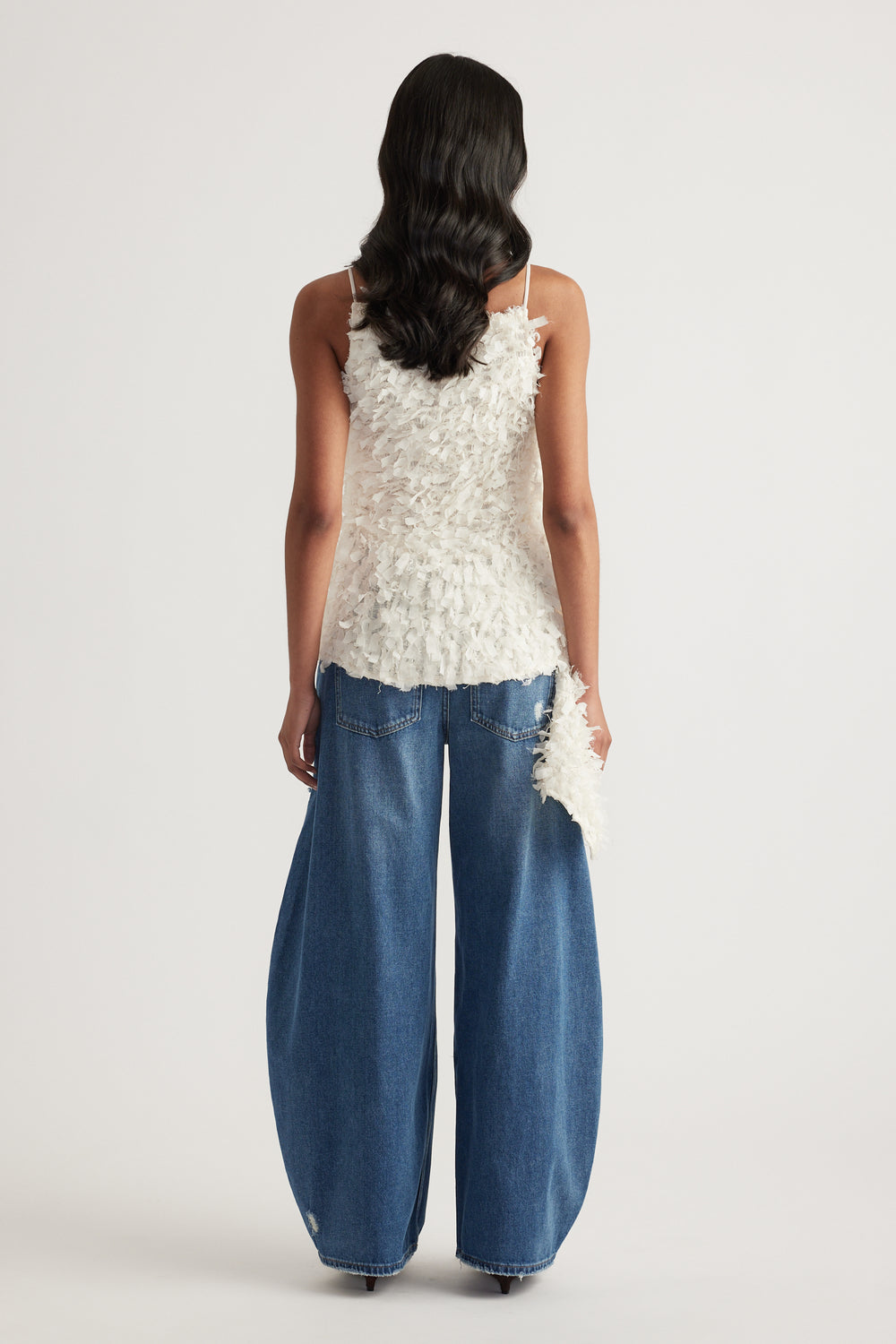 Back view of a woman wearing a white textured sleeveless top with an asymmetric hem and wide leg blue jeans