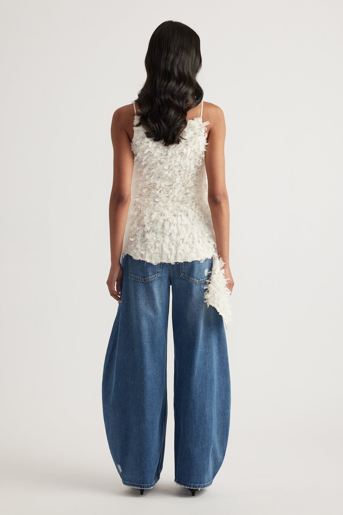 Back view of a woman wearing a white textured sleeveless top with an asymmetric hem and wide leg blue jeans