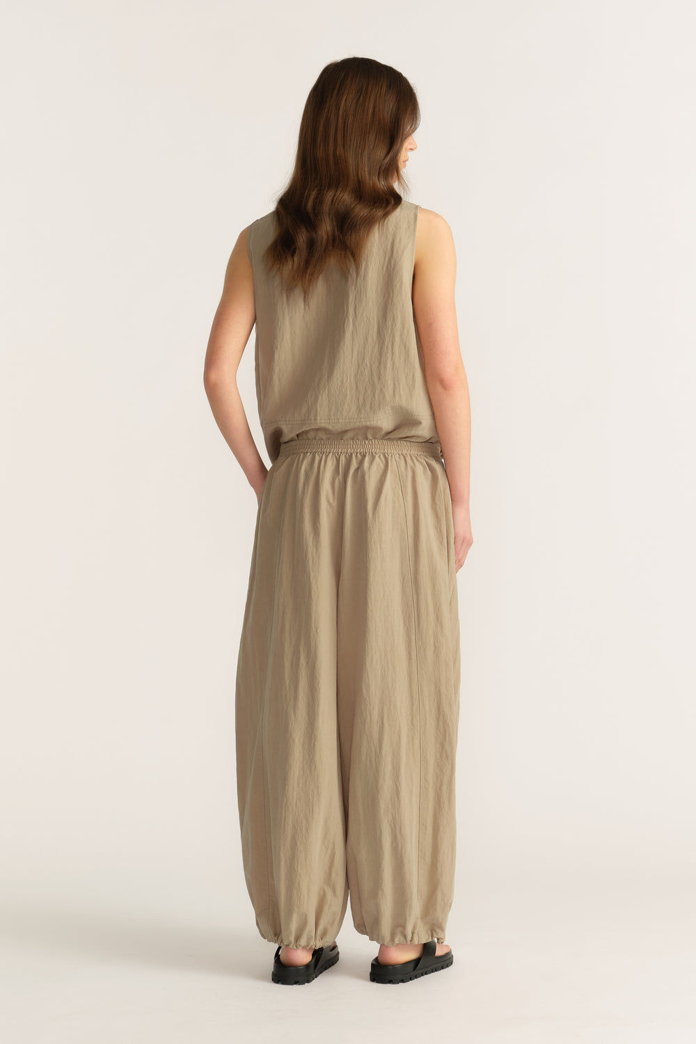 Back view of a woman wearing beige sleeveless top and relaxed balloon pants with elastic waist and black sandals