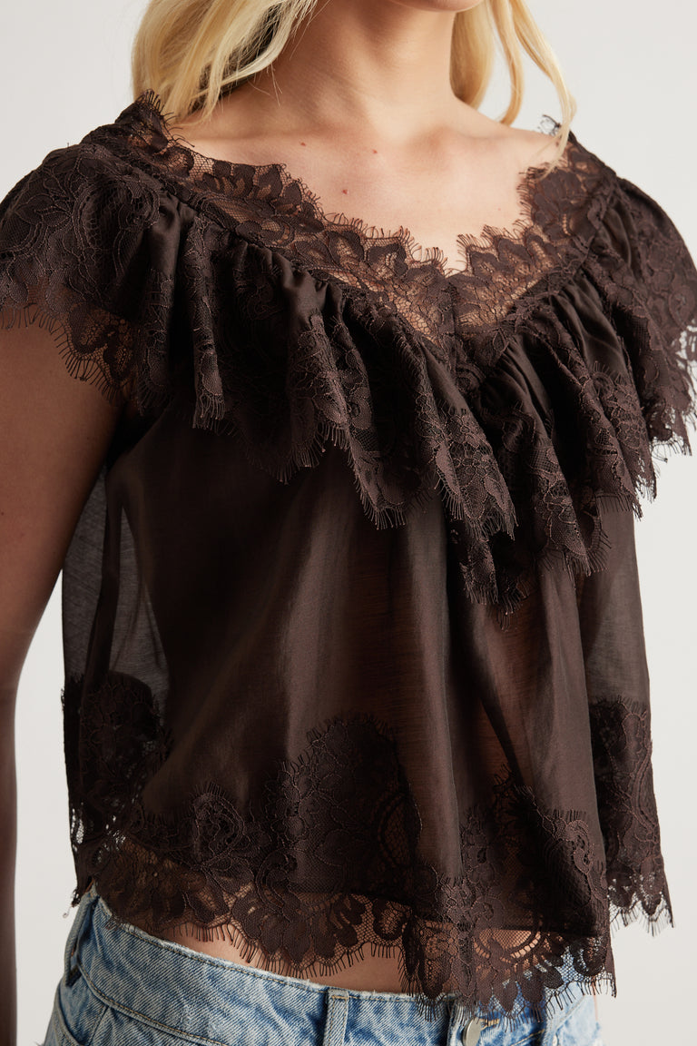 Woman wearing a dark brown lace top with ruffle detailing, flutter sleeves and a scalloped hem