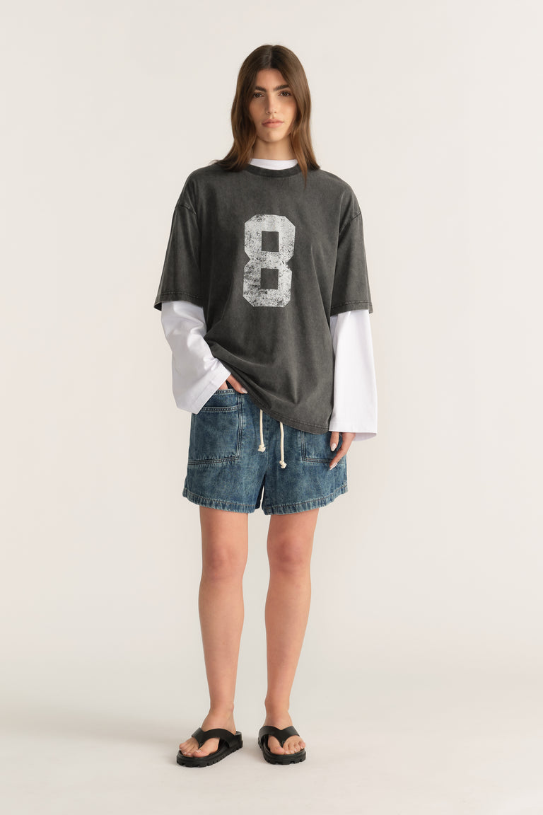 Woman wearing an oversized washed charcoal tee with an 8 graphic, white layered long sleeves and denim shorts