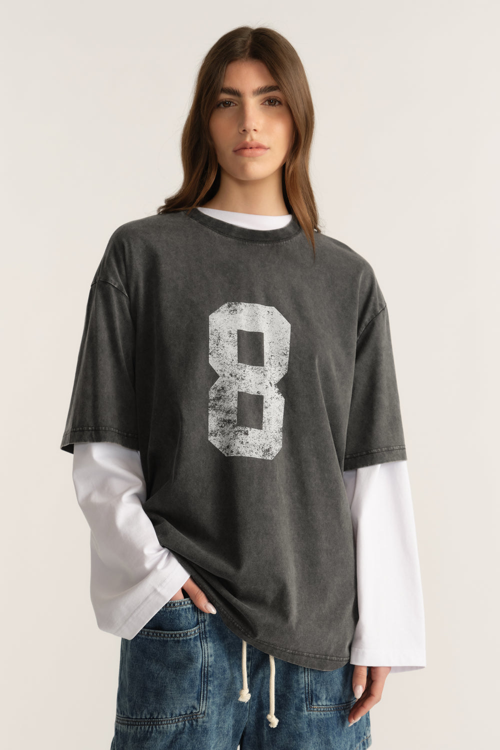 Woman wearing an oversized washed charcoal tee with a distressed number graphic, layered over a white long sleeve and jeans