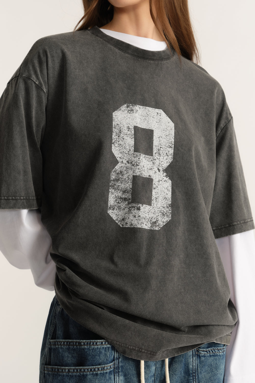 Woman wearing an oversized washed charcoal tee with a distressed number graphic over a white long sleeve top and jeans