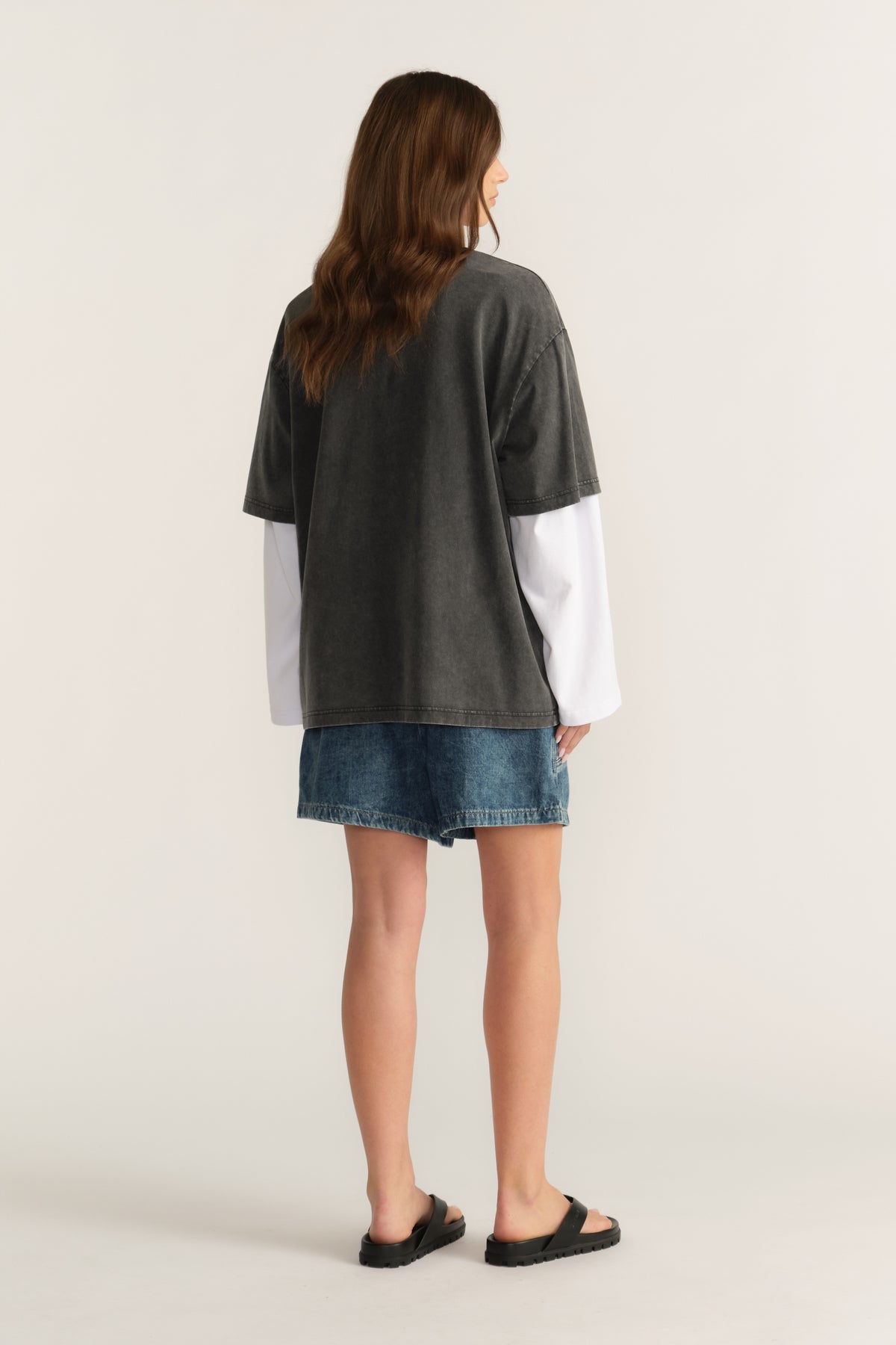 Back view of woman in an oversized charcoal tee layered over a white long sleeve top with denim shorts and sandals