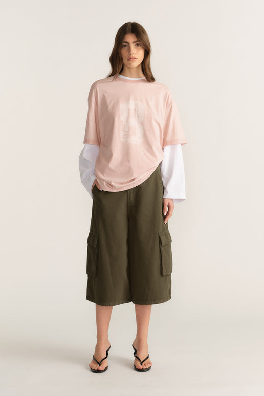 Model in a pink oversized tee layered over a white long sleeve top with olive cargo shorts and flip flops