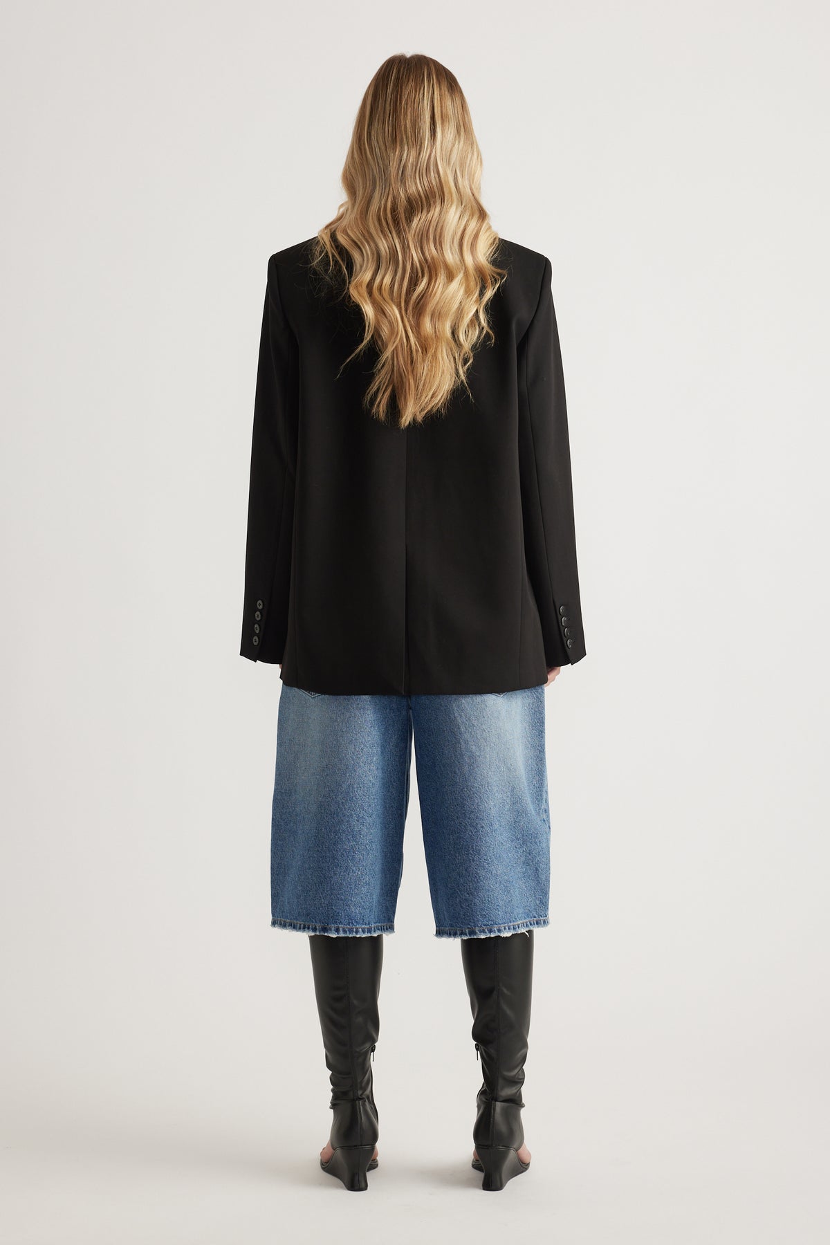 Back view of woman wearing an oversized black blazer with blue wide leg jeans and knee high boots
