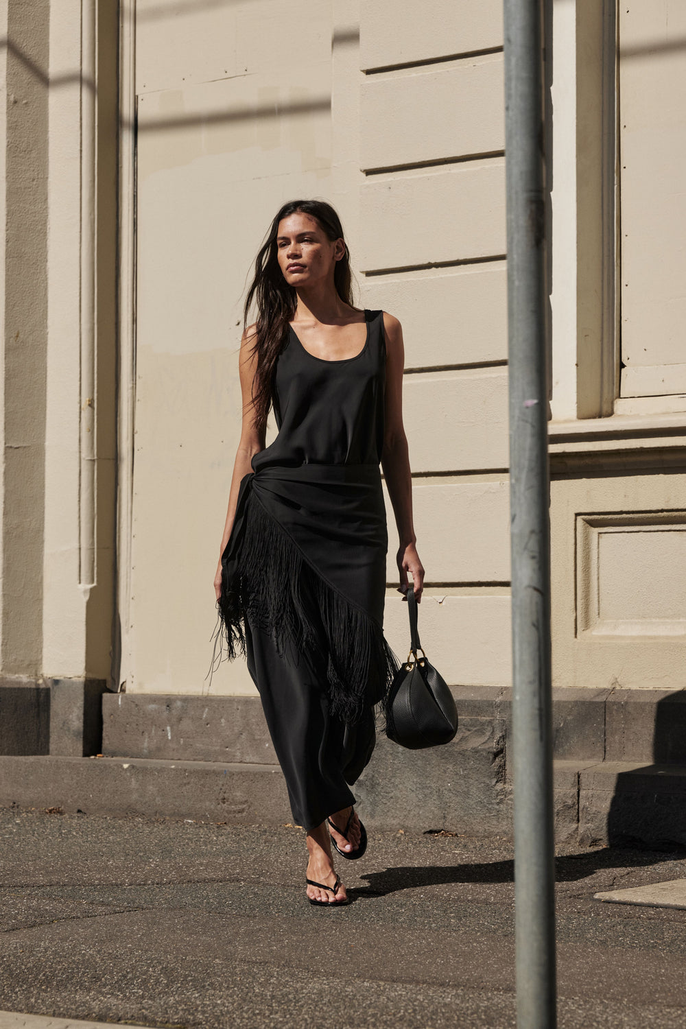 Woman wearing a black sleeveless maxi dress with fringe waist detail and carrying a handbag